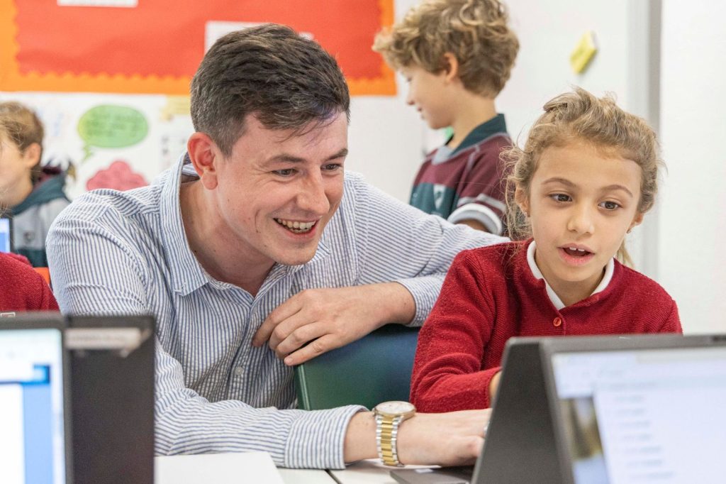 Profesor sonriendo mientras ayuda a una alumna que trabaja con un portátil en clase en Dallington School